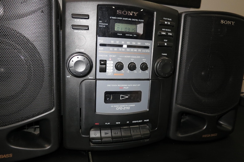 May include: Black Sony boombox with two speakers. The boombox has a digital display, a cassette player, and radio controls. The speakers have a mesh grill. The model number is CFD-Z110.