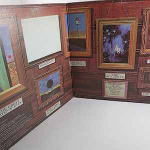 Puede incluir: Un libro abierto con obras de arte enmarcadas en una pared con paneles de madera. El arte incluye pinturas abstractas y paisajes, con t&iacute;tulos como "The Great Gates of Kiev" y "The Old Castle". El libro est&aacute; abierto, mostrando varias im&aacute;genes enmarcadas.