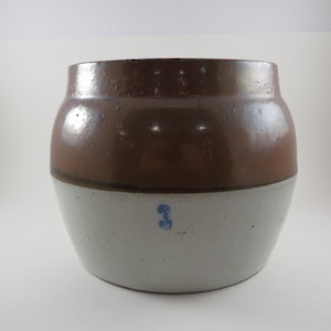 May include: A two-tone stoneware crock with a brown upper section and a gray lower section. The crock has a rounded shape and a small blue number 3 painted on the front. The surface has a glossy finish.