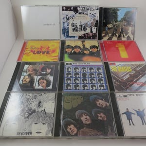 May include: A collection of The Beatles CDs, featuring album covers with various designs and colors. The CDs are in their original jewel cases, showcasing iconic album art and band photos. The collection includes albums like "Love," "1," and "Revolver."