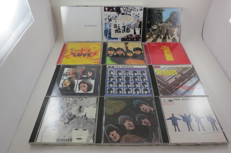 May include: A collection of The Beatles CDs, featuring album covers with various designs and colors. The CDs are in their original jewel cases, showcasing iconic album art and band photos. The collection includes albums like "Love," "1," and "Revolver."