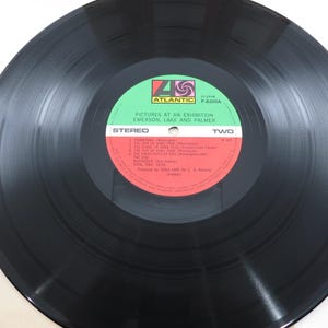 Puede incluir: Un disco de vinilo negro con una etiqueta verde y roja. La etiqueta dice "PICTURES AT AN EXHIBITION EMERSON, LAKE AND PALMER" y "STEREO TWO". El disco est&aacute; sobre una superficie blanca.