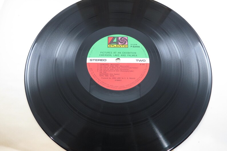 Puede incluir: Un disco de vinilo negro con una etiqueta verde y roja. La etiqueta dice "PICTURES AT AN EXHIBITION EMERSON, LAKE AND PALMER" y "STEREO TWO". El disco est&aacute; sobre una superficie blanca.