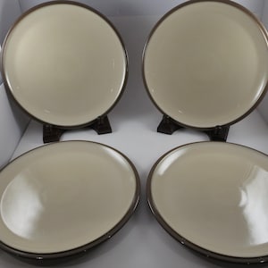 May include: Four cream-colored ceramic plates with a dark brown rim. Two plates are upright on small stands, while the other two are flat. The plates have a glossy finish and are arranged in a square formation.