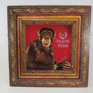 May include: A framed album cover featuring a chimpanzee holding a flower. The album cover has a red background with the words "Naked Talking Heads" in white text. The frame is brown with gold accents.