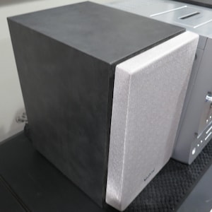 May include: A black and gray speaker with a silver audio component. The speaker has a textured gray front and a black top. The audio component has knobs and buttons.