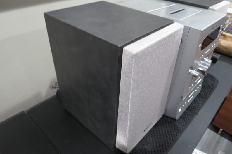 May include: A black and gray speaker with a silver audio component. The speaker has a textured gray front and a black top. The audio component has knobs and buttons.