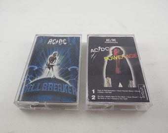 May include: Two cassette tapes with AC/DC album covers. The first tape is "Highway to Hell" with a blue and black cover featuring a man in a leather jacket. The second tape is "Powerage" with a red, yellow, and black cover featuring a man in a leather jacket.