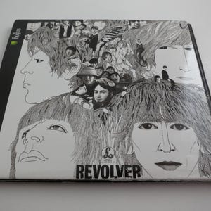 May include: A black and white album cover featuring a detailed illustration of multiple faces. The word "REVOLVER" is printed in bold, black letters at the bottom. The Beatles logo is visible in the upper left corner.