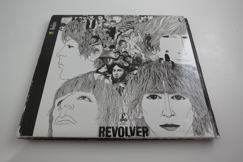 May include: A black and white album cover featuring a detailed illustration of multiple faces. The word "REVOLVER" is printed in bold, black letters at the bottom. The Beatles logo is visible in the upper left corner.