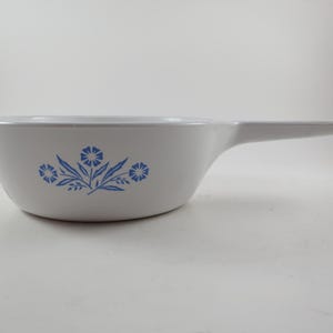 May include: White casserole dish with a handle, featuring a blue floral design. The dish has a rounded shape and a handle extending from one side. A classic piece for serving and baking.