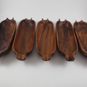 May include: Five wooden serving bowls with a dark brown finish. Each bowl has a long, oval shape with a rounded base and a small, raised handle at each end. The bowls are arranged in a row.