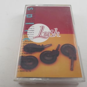 May include: A cassette tape case with a red and yellow cover featuring the band name "Lush" in a stylized font. The cover also displays four dark brown casters.