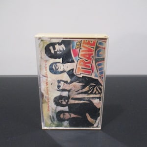 May include: A vintage cassette tape with a colorful image of a band called "The Traveling Wilburys" on the front. The band members are all wearing sunglasses and have their hair styled in a rock and roll fashion.