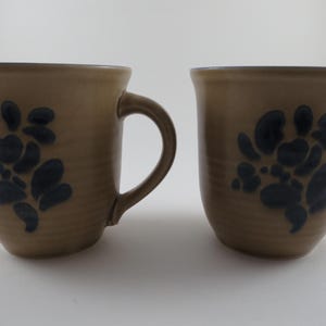 May include: Two beige ceramic mugs with dark blue floral designs. Each mug has a rounded body, a handle, and a dark blue rim. The mugs are displayed on a white surface.