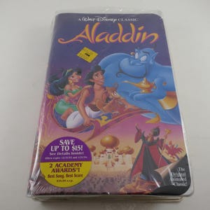 May include: Sealed VHS tape of the Disney animated classic, Aladdin. The cover features Aladdin, Jasmine, and the Genie on a magic carpet, with a purple and pink background. Includes text: "A Walt Disney Classic Aladdin" and "2 Academy Awards!"