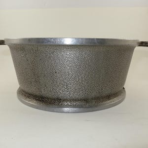 May include: A silver-colored metal bowl with a textured, dimpled surface. The bowl has a wide, rounded base and two small handles on either side. The rim of the bowl is smooth and shiny.