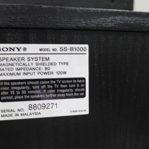 May include: A black wooden speaker system with a white rectangular label featuring the Sony logo, model number SS-B1000, 8 ohm impedance, 120W maximum input power, serial number 8809271, and text regarding magnetic shielding and TV screen interference.