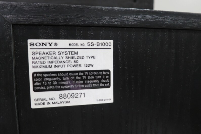 May include: A black wooden speaker system with a white rectangular label featuring the Sony logo, model number SS-B1000, 8 ohm impedance, 120W maximum input power, serial number 8809271, and text regarding magnetic shielding and TV screen interference.