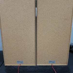 May include: Two rectangular speakers with a light brown, textured finish. Red wires extend from the bottom of each speaker. The speakers are positioned side by side, suggesting a stereo pair. The image is taken from the back of the speakers.