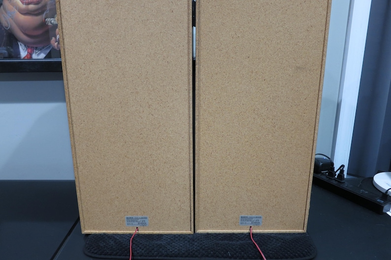 May include: Two rectangular speakers with a light brown, textured finish. Red wires extend from the bottom of each speaker. The speakers are positioned side by side, suggesting a stereo pair. The image is taken from the back of the speakers.