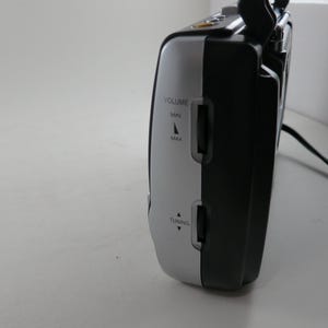 May include: A black and silver portable radio with volume and tuning controls. The radio has a handle and a power cord. The side of the radio features the words "VOLUME" and "TUNING" with directional arrows.