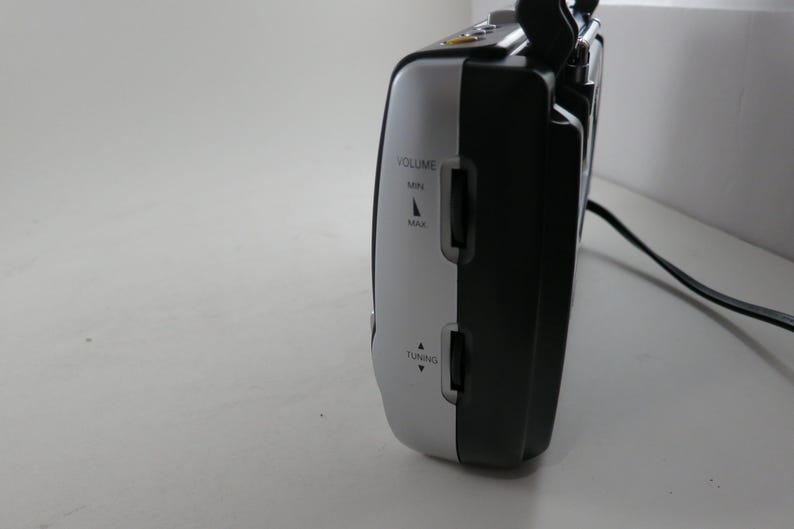 May include: A black and silver portable radio with volume and tuning controls. The radio has a handle and a power cord. The side of the radio features the words "VOLUME" and "TUNING" with directional arrows.