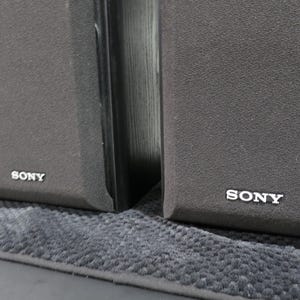 May include: Two black rectangular Sony speakers featuring dark grey fabric grilles covering the front panels. Each speaker has the Sony brand name printed in white lettering at the bottom center of the front grille, indicating the audio equipment.