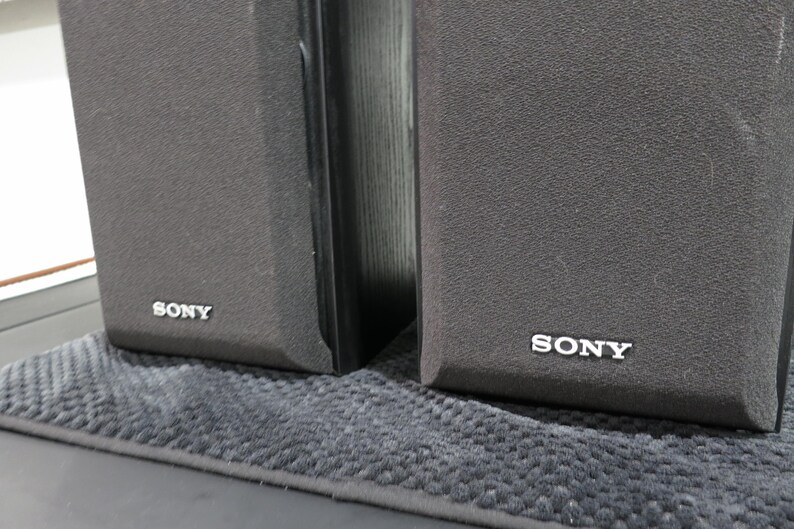 May include: Two black rectangular Sony speakers featuring dark grey fabric grilles covering the front panels. Each speaker has the Sony brand name printed in white lettering at the bottom center of the front grille, indicating the audio equipment.