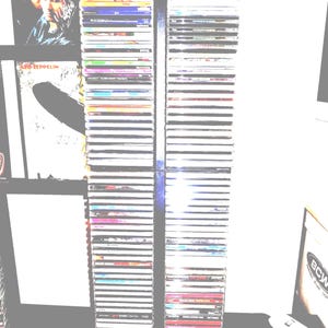 May include: Two tall, vertical stacks of compact disc cases arranged in a dark metal storage rack. The spines of the CDs display various colors, text, and graphic designs. To the left, a framed poster features a black and white image of a band.