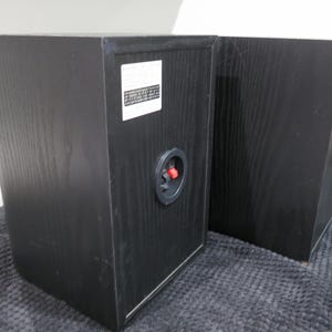 May include: Two black wooden bookshelf speakers shown from the rear. Each speaker features a white rectangular label with printed text and a circular input terminal with a red and black spring-loaded clip for connecting audio speaker wires.