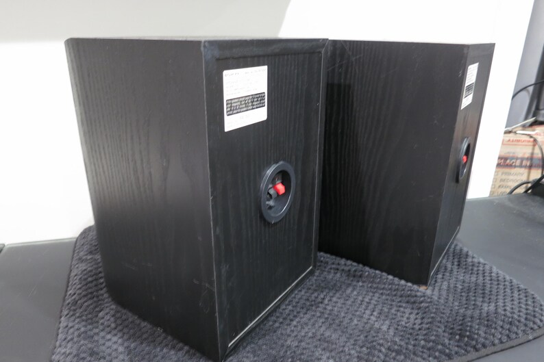 May include: Two black wooden bookshelf speakers shown from the rear. Each speaker features a white rectangular label with printed text and a circular input terminal with a red and black spring-loaded clip for connecting audio speaker wires.