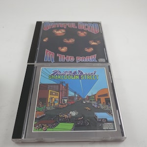 May include: Two Grateful Dead CD albums. The top album is "In the Dark" with a black cover and the band's name in red. The bottom album is "Shakedown Street" with a street scene illustration.