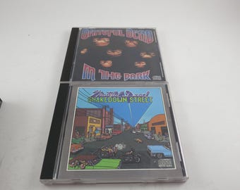 May include: Two Grateful Dead music CDs in plastic jewel cases. The top case features the In the Dark album cover with a black background and floating faces. The bottom case shows the Shakedown Street album cover with a colorful street scene.