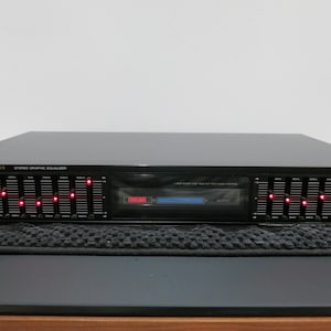 May include: A black Fisher Studio Standard stereo graphic equalizer featuring a horizontal row of vertical sliders with illuminated red indicators, a power button, tape monitor switch, and a central display panel with red and blue light accents.
