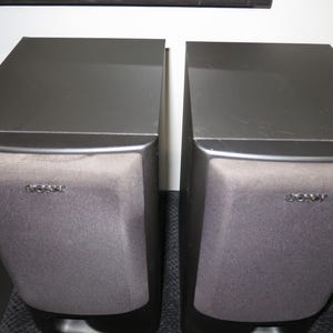 May include: A pair of black Sony bookshelf speakers with rectangular cabinets and dark grey fabric grilles covering the front panels. Each speaker features the Sony brand name printed in silver lettering on the upper center of the grille.