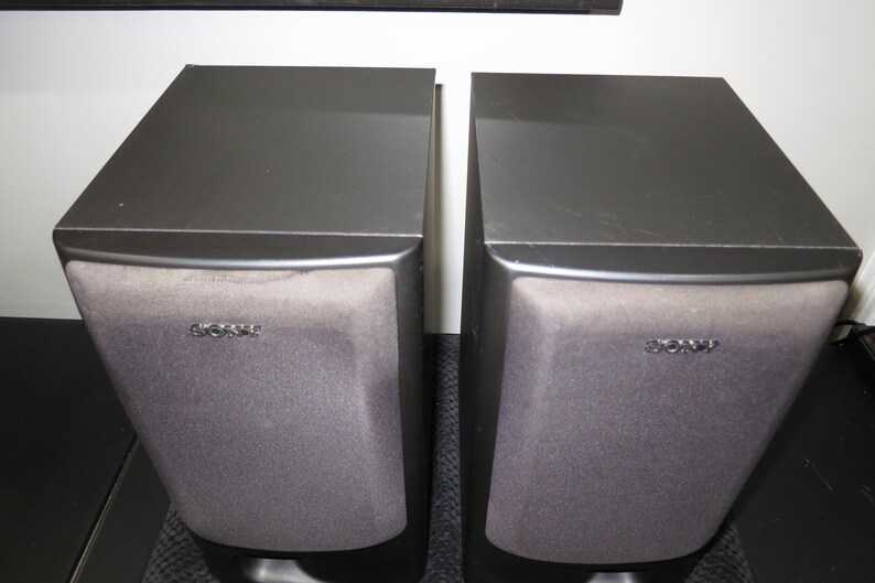 May include: A pair of black Sony bookshelf speakers with rectangular cabinets and dark grey fabric grilles covering the front panels. Each speaker features the Sony brand name printed in silver lettering on the upper center of the grille.