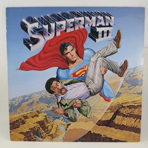 May include: Superman III album cover featuring Superman in a blue suit and red cape carrying a man in a suit over a canyon. The album title is in a stylized font with lightning bolts.
