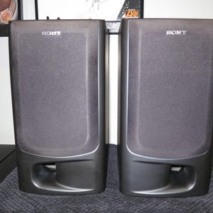 May include: A pair of black Sony bookshelf speakers with rectangular fabric grilles covering the main drivers and a horizontal bass reflex port located at the bottom of each unit. The Sony brand name is printed in white at the top center.