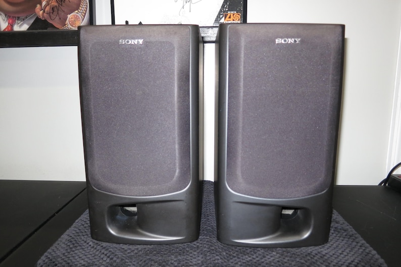 May include: A pair of black Sony bookshelf speakers with rectangular fabric grilles covering the main drivers and a horizontal bass reflex port located at the bottom of each unit. The Sony brand name is printed in white at the top center.