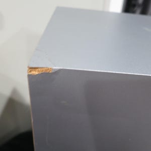 以下が含まれることがあります： 滑らかなマット仕上げのグレーの長方形スピーカーキャビネット。左上の角には目に見える損傷があり、グレーの表面コーティングが剥がれて、外層の下にある明るい茶色の木材のような素材が露出しています。
