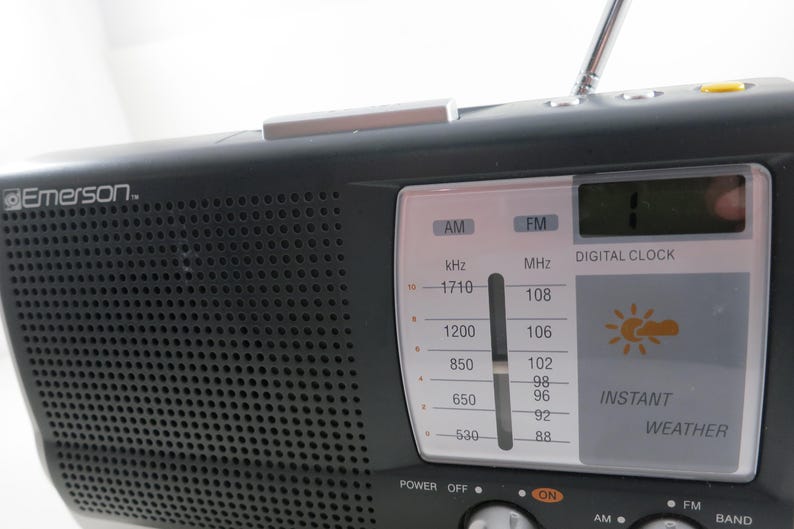 May include: Black Emerson radio with a digital clock and instant weather display. The radio has an AM/FM tuner, a telescoping antenna, and a speaker grill. The display shows the time and weather icons.