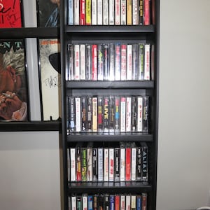 May include: A tall, black, five-shelf wooden media storage tower filled with various music cassette tapes. Each shelf is packed with rows of plastic cassette cases featuring colorful spine labels with artist names and album titles in various fonts.