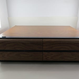 May include: A rectangular wooden cabinet with four drawers, featuring a dark wood grain finish. The top surface is a lighter wood tone, and the edges are accented with black trim. The cabinet is set against a white background.