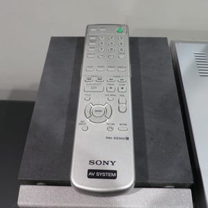 May include: Silver Sony RM-SS300 AV System remote control with numerous buttons, including volume, channel, and menu controls. The remote is on a dark gray surface, with a silver AV system component visible to the right.