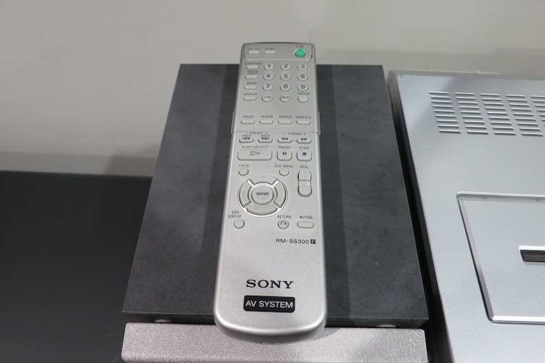 May include: Silver Sony RM-SS300 AV System remote control with numerous buttons, including volume, channel, and menu controls. The remote is on a dark gray surface, with a silver AV system component visible to the right.