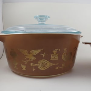 May include: A brown casserole dish with a clear glass lid, featuring gold-colored illustrations of an eagle, a rooster, a coffee grinder, and a fireplace bellows. The round ceramic pot has a side handle and a smooth, matte-finish exterior surface.