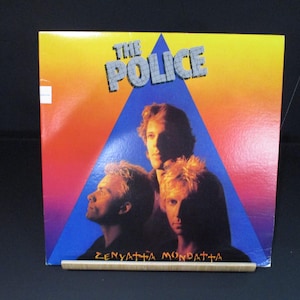 May include: A vinyl record album cover featuring a band called "The Police". The cover is a bright orange, yellow, and blue color scheme with a triangle shape in the center. The band members are pictured in the triangle, with the band's name in large white letters at the top. The album title is "Zenyatta Mondatta" in red letters at the bottom.