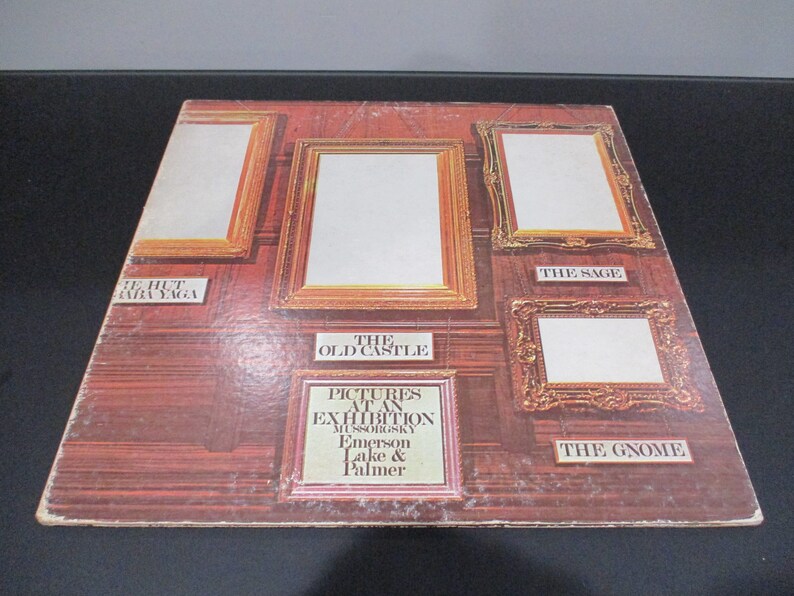 Op de afbeelding: Een vintage vinyl platenhoes met een bruine houten muur met vijf ingelijste foto's. Het album is getiteld "Pictures at an Exhibition" van Mussorgsky, Emerson Lake & Palmer. De titels van de foto's zijn "The Hut on Fowl's Legs", "The Old Castle", "The Sage" en "The Gnome".