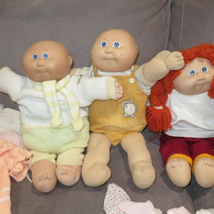 May include: Three dolls with round faces and blue eyes. The left doll wears a white sweater with a yellow scarf and light yellow pants. The center doll wears a yellow overall with an elephant graphic. The right doll has red yarn hair.
